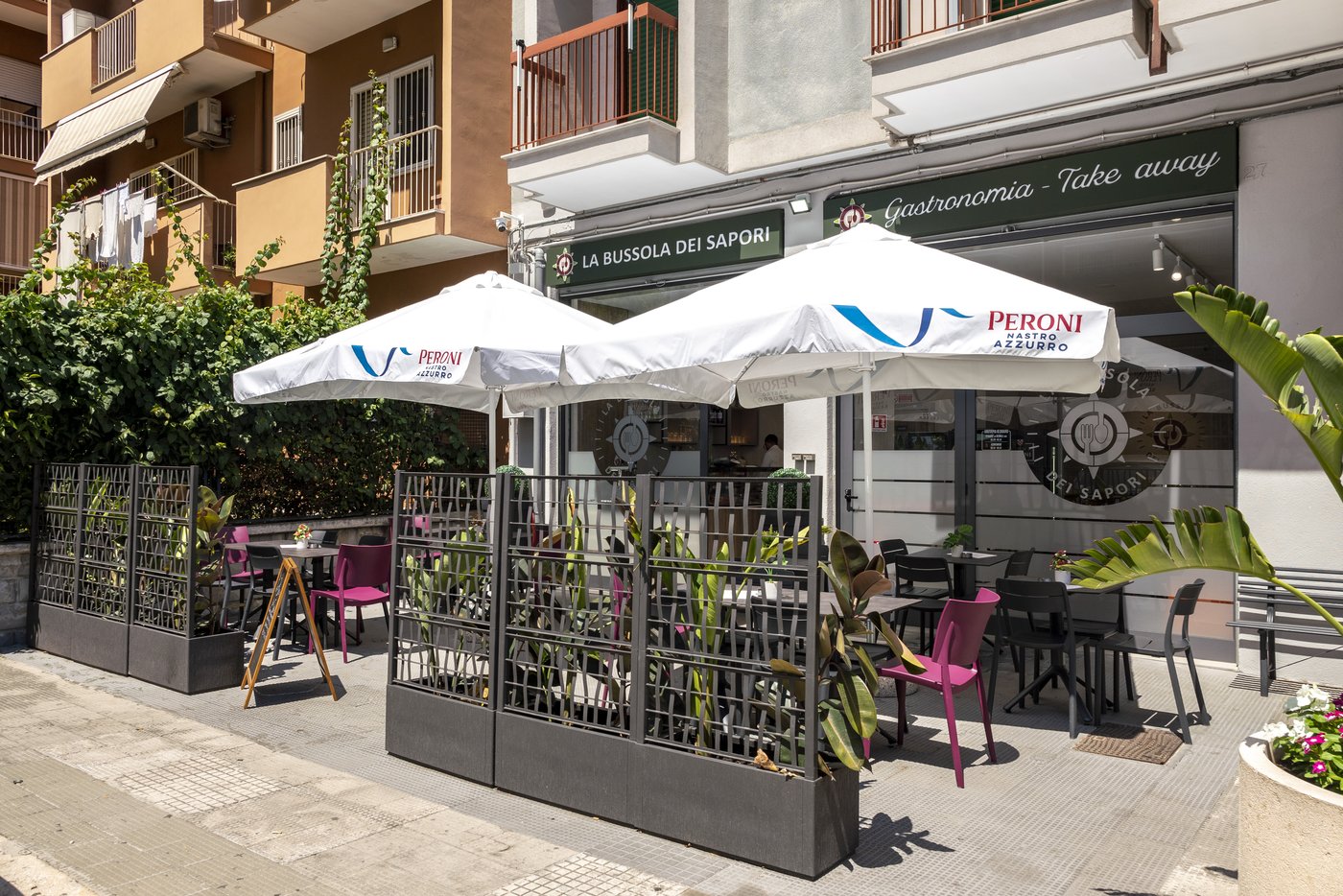 Dehors de La Bussola dei Sapori: ombrelloni Peroni, sedie fucsia, fioriere grigie e insegna Gastronomia - Take away