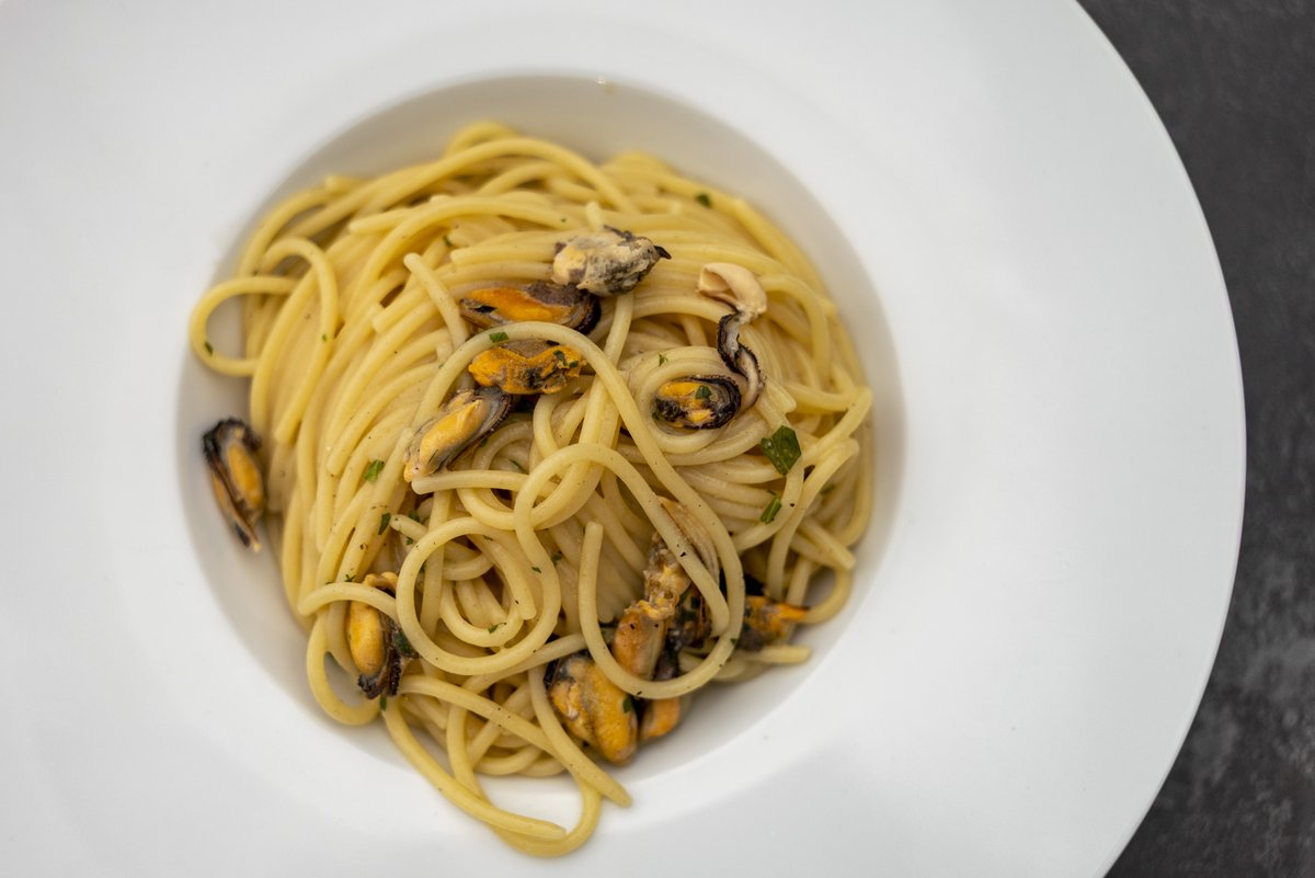 Spaghetti con cozze sgusciate e prezzemolo tritato in un piatto fondo bianco su un piano scuro, La Bussola dei Sapori